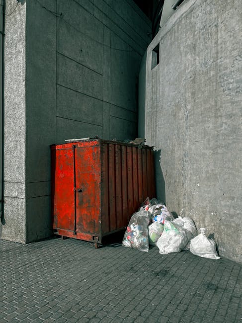 A large, rusty red metal container, used for rubbish disposal, positioned against a dark green wall and a light grey concrete wall in a narrow urban alleyway. The container has a weathered appearance with visible scratches, rust patches, and a black padlock on the front. Several clear plastic bags filled with various waste items, including visible plastic bottles and packaging, are placed on the ground beside the container, some leaning against it. The ground is paved with dark grey interlocking bricks, and the environment is illuminated by natural daylight, creating a neutral, functional setting typical for private waste collection areas or independent disposal points. The surrounding walls are smooth, with the building surface on the left being dark green with horizontal paneling, while the wall on the right is a plain light grey concrete. This scene exemplifies an accessible, on-site waste storage solution, possibly managed separately from official municipal rubbish collection services, aligning with alternative rubbish handling methods.