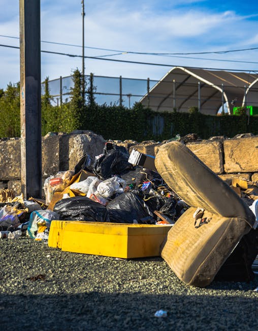 A close-up view of an unmanaged pile of mixed rubbish and waste materials positioned on a paved surface, consisting of several black plastic garbage bags, some partially torn open revealing paper waste, and a yellow plastic container lying on its side. Next to the pile, a discarded old beige car tire leans against a stone wall constructed from large, uneven concrete blocks. In the background, a metal utility pole with power lines extends upward, and behind it, a green hedge and a large, semi-circular fabric-covered structure are visible. The sky is partly cloudy with patches of blue, suggesting daylight, and the scene reflects an environment where waste has accumulated outdoors, indicative of independent waste collection or non-standard rubbish removal services, such as those offered by Waste Collection Bayswater.