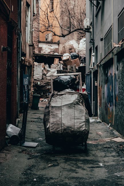 A large, rusty red metal container, used for rubbish disposal, positioned against a dark green wall and a light grey concrete wall in a narrow urban alleyway. The container has a weathered appearance with visible scratches, rust patches, and a black padlock on the front. Several clear plastic bags filled with various waste items, including visible plastic bottles and packaging, are placed on the ground beside the container, some leaning against it. The ground is paved with dark grey interlocking bricks, and the environment is illuminated by natural daylight, creating a neutral, functional setting typical for private waste collection areas or independent disposal points. The surrounding walls are smooth, with the building surface on the left being dark green with horizontal paneling, while the wall on the right is a plain light grey concrete. This scene exemplifies an accessible, on-site waste storage solution, possibly managed separately from official municipal rubbish collection services, aligning with alternative rubbish handling methods.