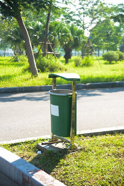A row of five large wheeled rubbish bins lined up against a plain, light-colored wall with a smooth surface, possibly painted concrete or plaster. Each bin is made of durable black plastic with a textured surface, equipped with small black wheels at the base for mobility, and features a bright yellow lid with a ribbed finish, contrasting with the dark body. The bins are positioned on a flat, gray paving surface, likely concrete, with slight dirt marks and wear visible around the wheels. White oval labels with printed text and barcode stickers are affixed to the front of each bin, indicating ownership or identification codes. The scene appears to be in an outdoor or semi-outdoor setting, possibly a service alley or disposal area used by Waste Collection Bayswater for daily waste management tasks or private rubbish removal services, supporting local waste disposal needs outside regular council collections.