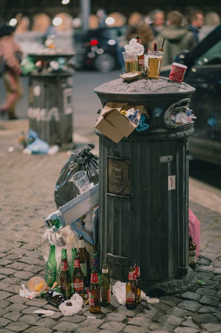 A black, traditional metal public trash bin with a rounded dome lid, situated on a cobblestone pavement in an urban outdoor setting. The bin's surface shows signs of weathering, with visible scratches and slight rust. The lid is topped with several disposable coffee cups, crumpled paper, and plastic bottles, suggesting recent use. On the side of the bin, there is a corrugated cardboard box with its edges frayed, containing miscellaneous rubbish. Surrounding the base on the ground are empty glass bottles in various colours, including green and brown, some smashed, alongside crumpled paper, plastic wrappers, and discarded food containers. In the background, out of focus, there are additional bins, parked cars, and pedestrians walking along the street, indicating a busy urban environment. Waste Collection Bayswater provides rubbish removal services for private waste management, and this scene exemplifies improper disposal often addressed through their on-site clearance solutions.