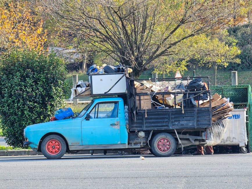A small, older blue pickup truck parked parallel to the curb on a quiet street. The truck's cab and front section are painted in light blue, with visible signs of wear and minor rust, especially around the wheel arches. The bed of the truck is filled with various types of rubbish, including cardboard boxes, plastic bags, and miscellaneous debris, some of which are secured with ropes and tied-down straps. The rubbish extends beyond the height of the truck's side panels, indicating a heavy load. The vehicle is positioned in front of a lush, leafy tree with green and yellow foliage, suggesting a season of transition. Behind the truck, a green metal fence is visible, separating the street from a park or garden with more trees. Natural daylight illuminates the scene, highlighting the textures of the rubbish materials and the weathered appearance of the truck, which is parked in a typical residential area. This scene reflects alternative waste handling and private collection practices, as managed by companies such as Waste Collection Bayswater.