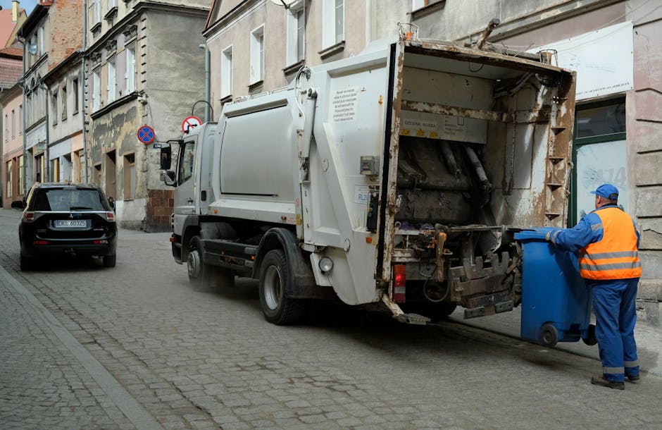 A waste collection vehicle, primarily white with visible signs of rust and wear on its rear compartment, is positioned on a narrow city street next to a row of old, multistory buildings constructed with stone and brick facades. The truck's rear loader mechanism is open, showing internal components used for compacting rubbish. A worker dressed in a blue uniform with an orange high-visibility vest and a blue cap is standing beside the vehicle, emptying a blue wheeled bin into the rear compartment. The street surface is cobblestone, and parked cars, including a black vehicle, are visible along the curb. Signage indicating parking restrictions can be seen attached to the buildings, and the overall environment appears to be a typical urban setting. This scene exemplifies independent rubbish collection activity outside traditional municipal waste services, highlighting private waste management practices familiar in a busy neighbourhood.