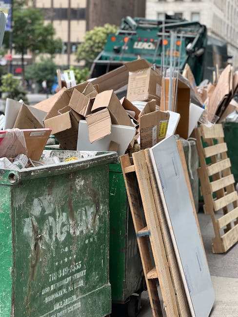 A small, older blue pickup truck parked parallel to the curb on a quiet street. The truck's cab and front section are painted in light blue, with visible signs of wear and minor rust, especially around the wheel arches. The bed of the truck is filled with various types of rubbish, including cardboard boxes, plastic bags, and miscellaneous debris, some of which are secured with ropes and tied-down straps. The rubbish extends beyond the height of the truck's side panels, indicating a heavy load. The vehicle is positioned in front of a lush, leafy tree with green and yellow foliage, suggesting a season of transition. Behind the truck, a green metal fence is visible, separating the street from a park or garden with more trees. Natural daylight illuminates the scene, highlighting the textures of the rubbish materials and the weathered appearance of the truck, which is parked in a typical residential area. This scene reflects alternative waste handling and private collection practices, as managed by companies such as Waste Collection Bayswater.