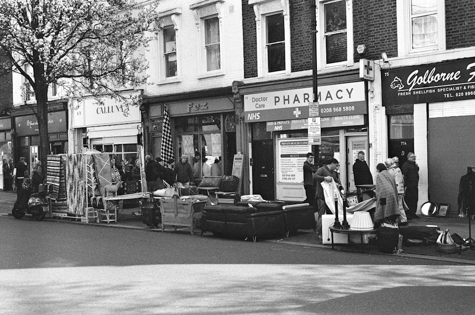 A street scene featuring a public outdoor market in front of a row of small shops, including a pharmacy and a seafood specialist store. Several people are gathered around a variety of stacked, discarded furniture and household items such as black sofas, wooden chairs, and small tables, which appear to have been temporarily placed on the sidewalk. The furniture surfaces have different textures, with some upholstered and others made of painted or polished wood, and they are arranged haphazardly across the pavement. A few vendors have set up makeshift stalls with fabric coverings and display racks for textiles or other small goods. The background shows the façades of brick buildings with windows, signage, and a tree partially obscuring some shop fronts. Natural light highlights the scene in a neutral tone, emphasizing the transient nature of the street’s clutter, which may relate to private or independent rubbish collection or on-site clearance undertaken by waste management services like Waste Collection Bayswater.