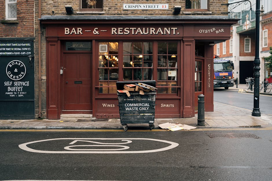 A black wheeled commercial waste bin with white text reading 'COMMERCIAL WASTE ONLY' is positioned directly in front of a brick-walled storefront of a pub or restaurant, which has a deep red wooden exterior and large glass windows displaying warm lighting inside. The bin is overflowing with flattened cardboard boxes and paper waste, some partially spilling onto the sidewalk. To the left of the bin, a small blackboard sign advertising a self-service buffet is mounted on the wall, and to the right, a short black bollard stands on the pavement. Behind the bin, adjacent to the building, there is a street with a black pole and several bicycles parked along the sidewalk. The surrounding environment includes a narrow street with a marked bus stop on the asphalt in the foreground and additional buildings visible in the background, indicating an urban setting suitable for private rubbish collection or waste management services by Waste Collection Bayswater, consistent with the context of alternative waste handling solutions in the area. Natural outdoor lighting highlights the textures of brickwork, wood, and litter.